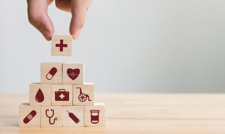 Das Bild zeigt eine Hand, die einen Holzklotz mit Kreuz auf einen Stapel mit Gesundheits- und Versicherungssymbolen setzt.