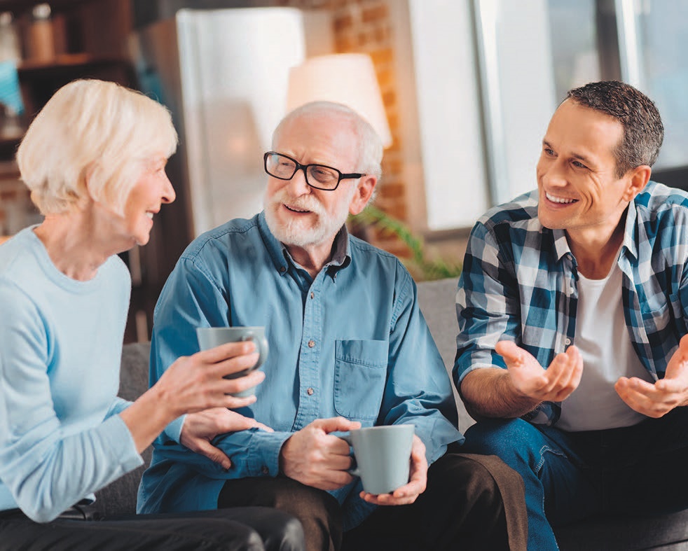 Das Bild zeigt drei Menschen unterschiedlicher Generationen, die entspannt zusammensitzen, Kaffee trinken und sich angeregt unterhalten.