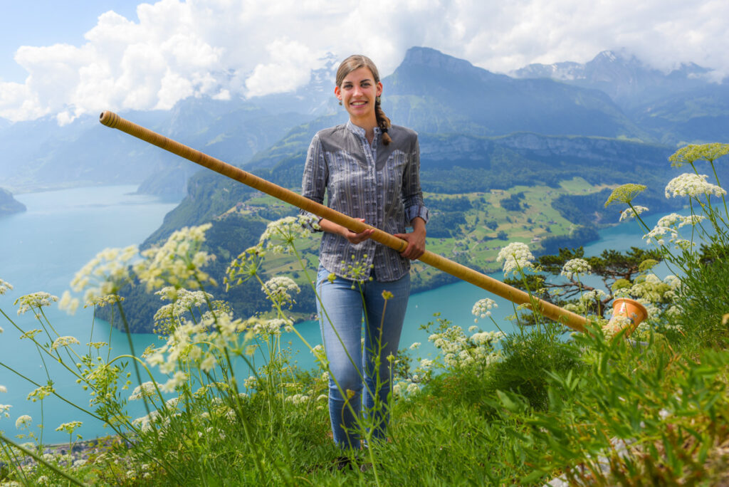 Das Bild zeigt eine junge Frau in Tracht, die auf einer Blumenwiese oberhalb eines Sees ein Alphorn hält, vor alpiner Bergkulisse.