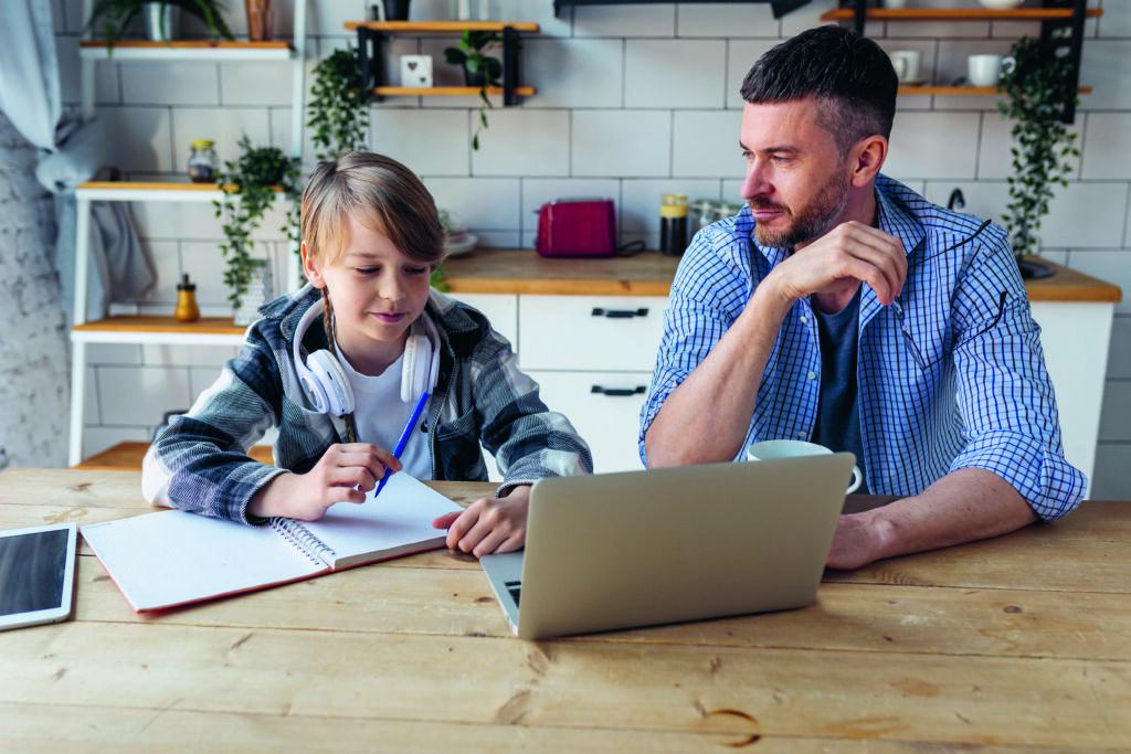 Vater hilft seinem Teenager-Sohn bei den Hausaufgaben, während er von zu Hause aus in der Küche arbeitet. Konzept von Elternschaft, Vaterschaft, gemeinsame Zeit verbringen. Einsatz von Technologie, Gadgets und Geräten zum Lernen.