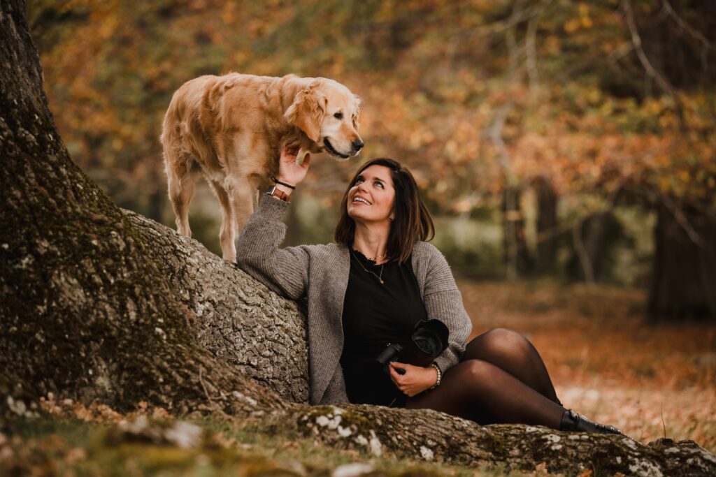 Frau sitzt im Wald angelehnt an einem Baustamm Hund staht auf dem Baumstamm